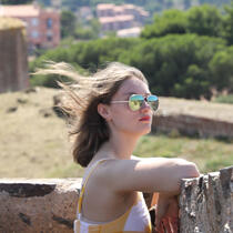 Person with shoulder length hair wearing sunglasses leaning on stone wall and looking right, facing wind, with trees behind them